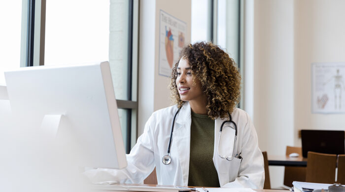 Female doctor sits in a brightly lit office at a desktop computer and responds to online reviews.