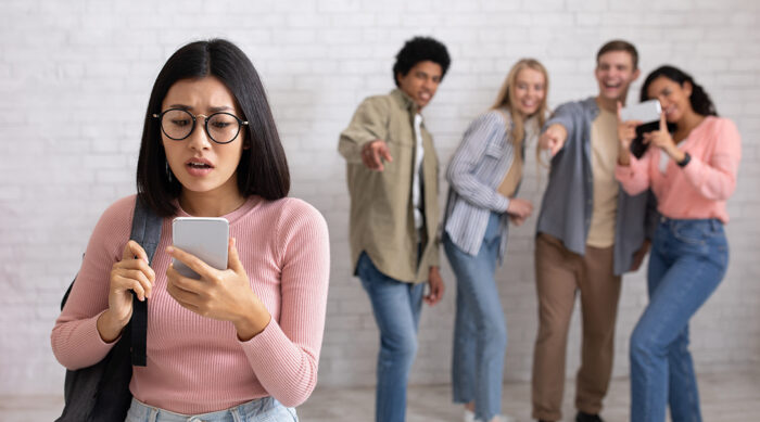 A group of teens point and laugh at a young girl who is being cyberbullied on social media.