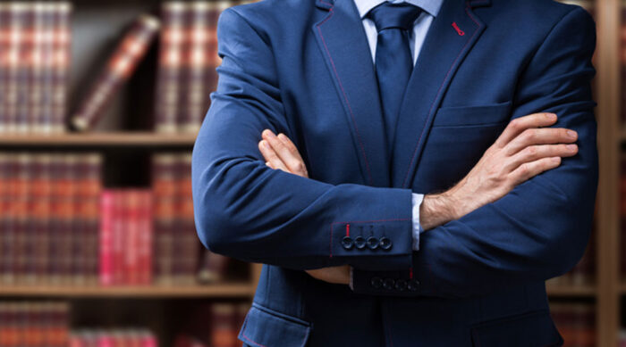 Midsection of a male lawyer wearing a navy suit with arms crossed in front of a bookcase.