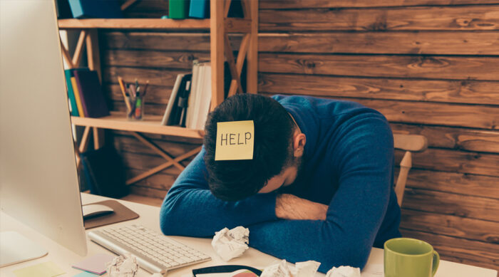 A young male business owner sits at a white desk with his head down asking to help.