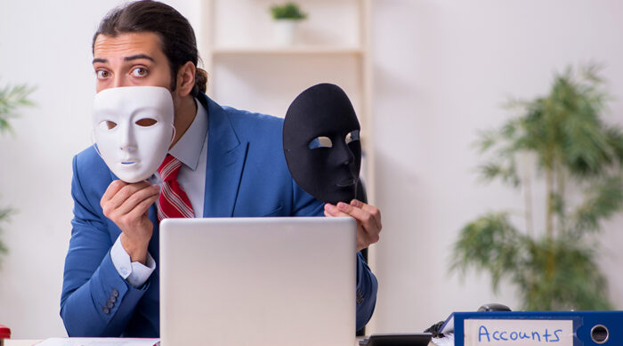 A man sits behind a computer and desk while hiding behind a black and white face covering mask.