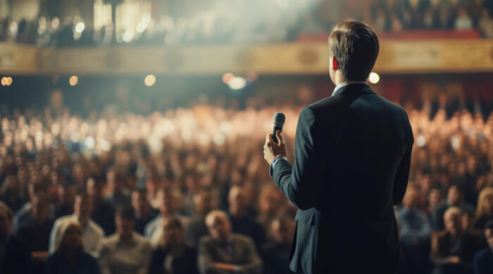 A man faces a large auditorium of people and publicly speaks.