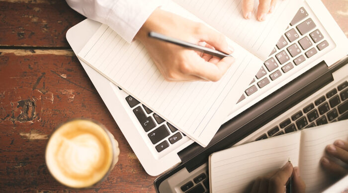 Woman holds a dark pen and writes in a light colored notebook near a laptop computer.