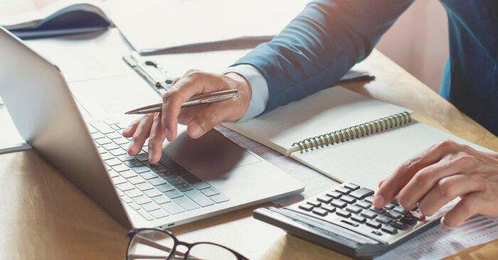 Tax attorney multitasking at a desk, using a laptop and calculator, surrounded by documents and a notepad, illustrating 501(c)(3) tax obligations.